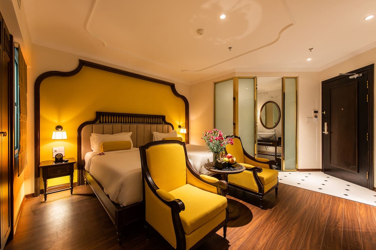 Habitación de hotel con una cama grande, dos sillones amarillos, una mesita con un jarrón de flores y un espejo en la pared.