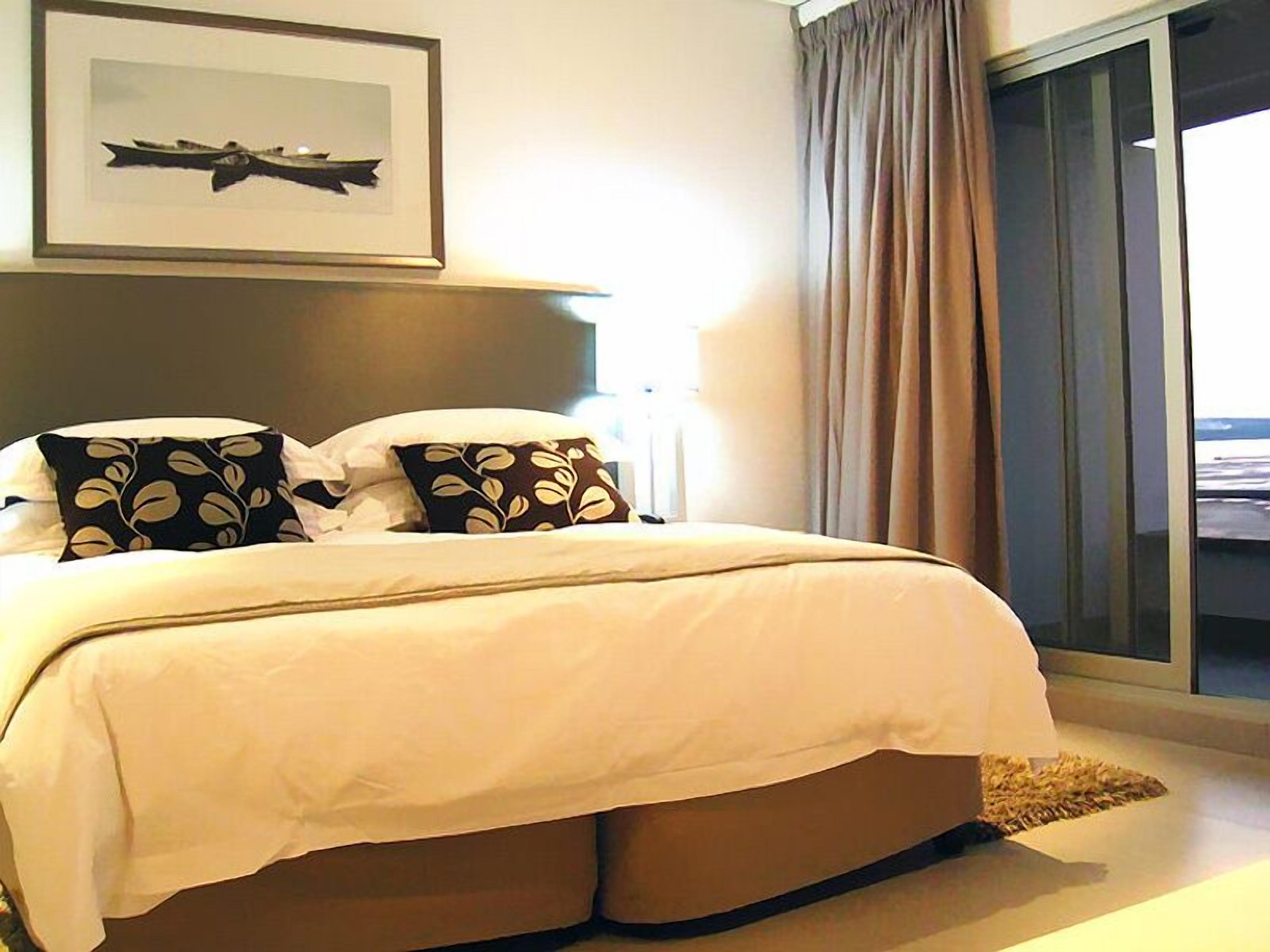 A neatly made bed with a white and beige duvet, two pillows with leaf patterns, a framed picture above the bed, and a view of the outside through a large window with curtains.