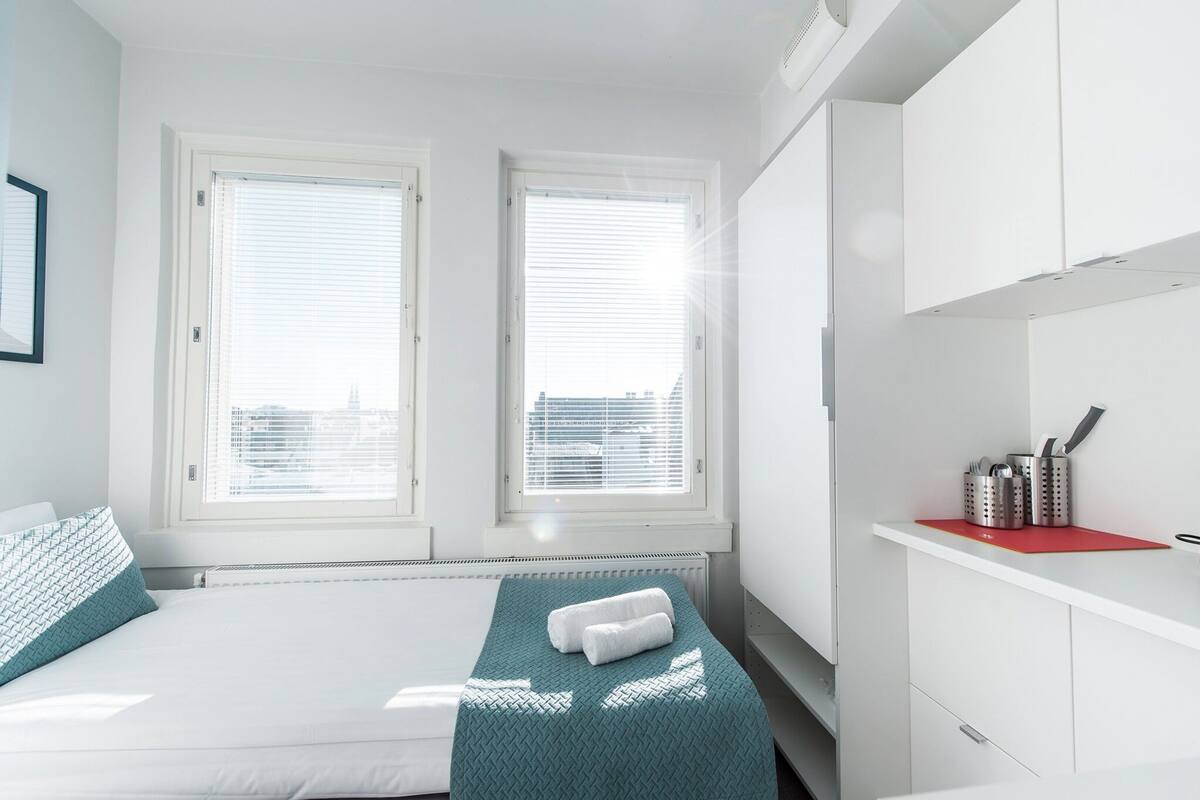 A modern bedroom with a bed, two windows with blinds, a radiator, and a kitchenette area with a red cutting board.