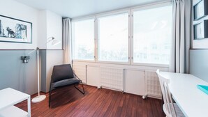 A room with wooden flooring, a white radiator, a black chair, a white table, and a large window with blinds. - Forenom Aparthotel Helsinki Kamppi (Helsinki)