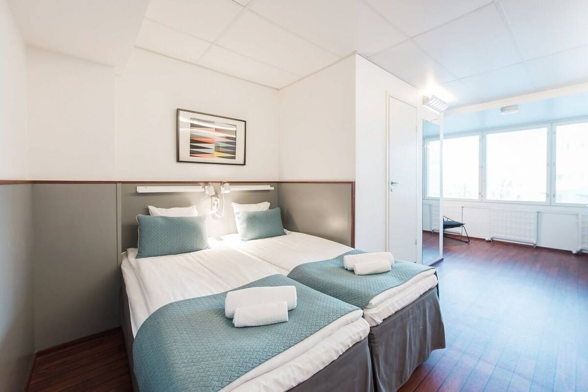 A hotel room with a double bed, two pillows, a bedside table, a framed picture, and a view of the outdoors through large windows.