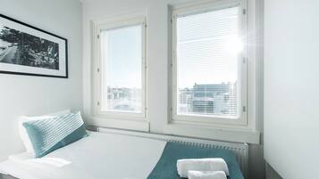 A modern hotel room with a bed, two windows with blinds, a radiator, and a framed picture on the wall.