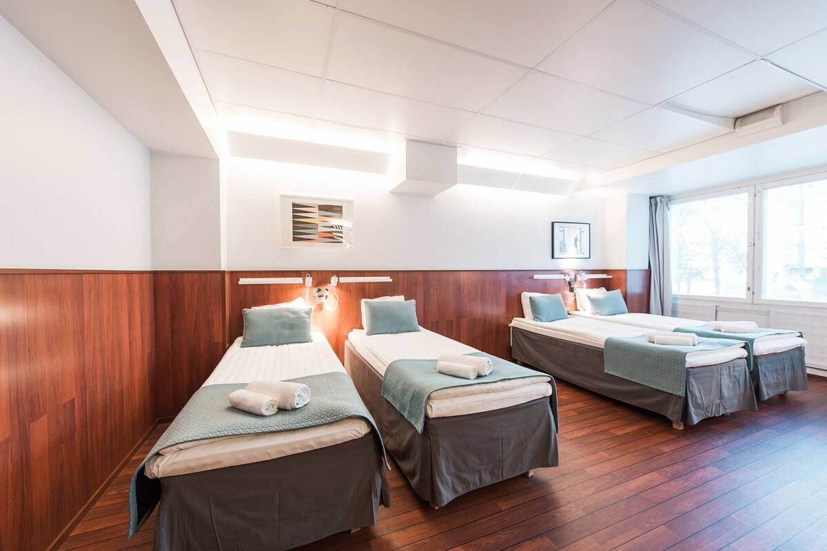 A hotel room with three single beds, wooden flooring, a large window, and a ceiling with recessed lighting.