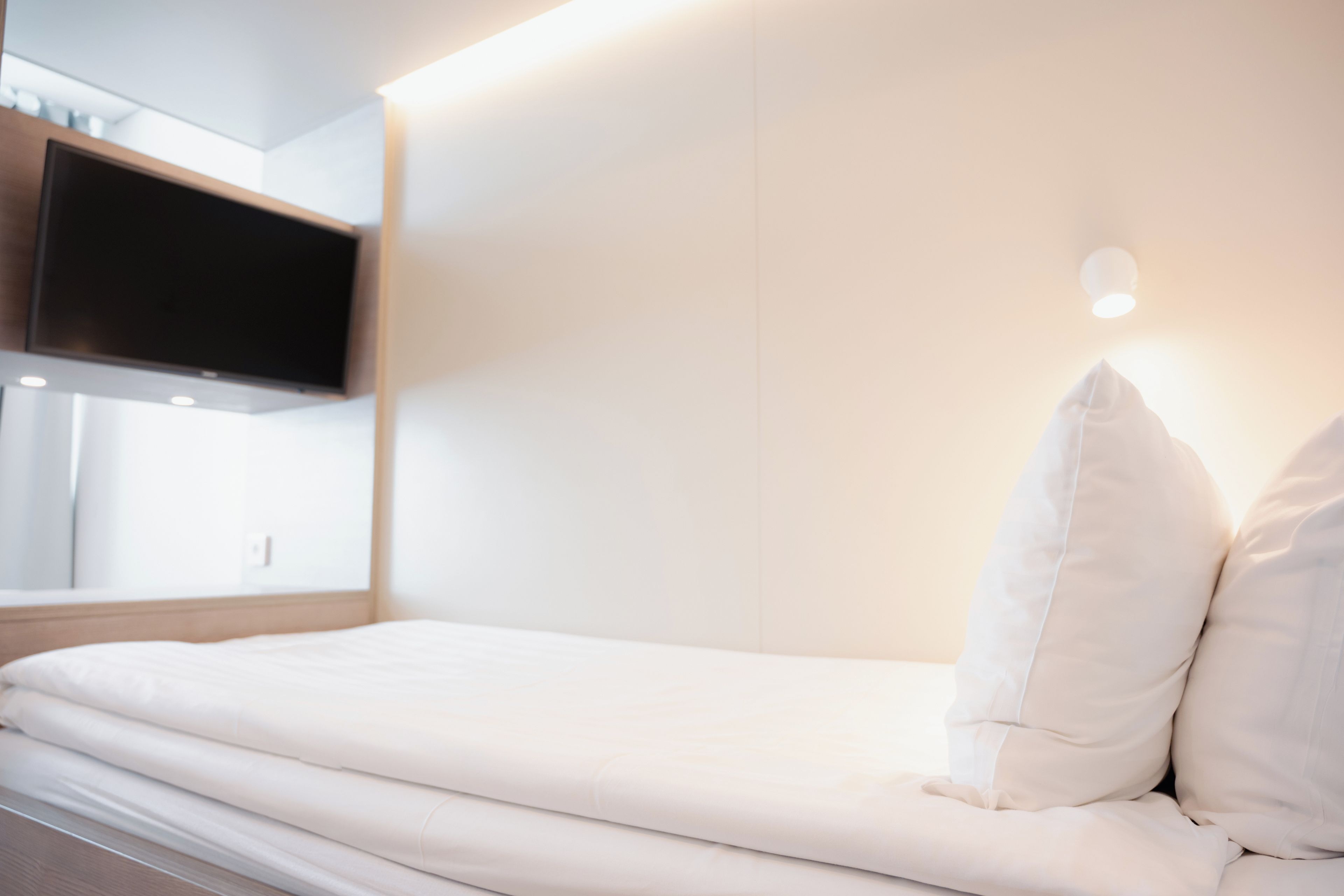 A neatly made bed with white bedding, a mounted television above it, and a wall-mounted light fixture.