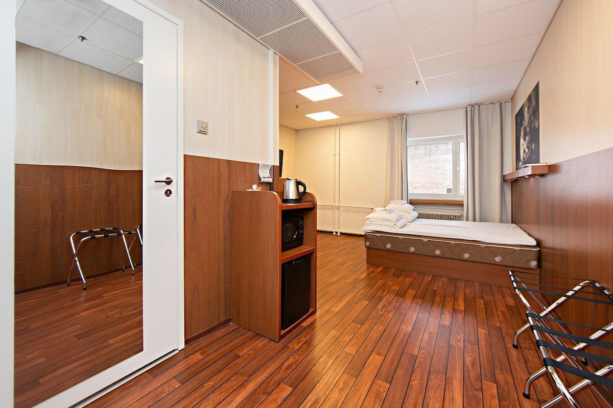 Une chambre d’hôtel de taille modeste, dotée d’un plancher en bois, d’un lit, d’une petite kitchenette, d’un radiateur et d’une fenêtre avec des rideaux.