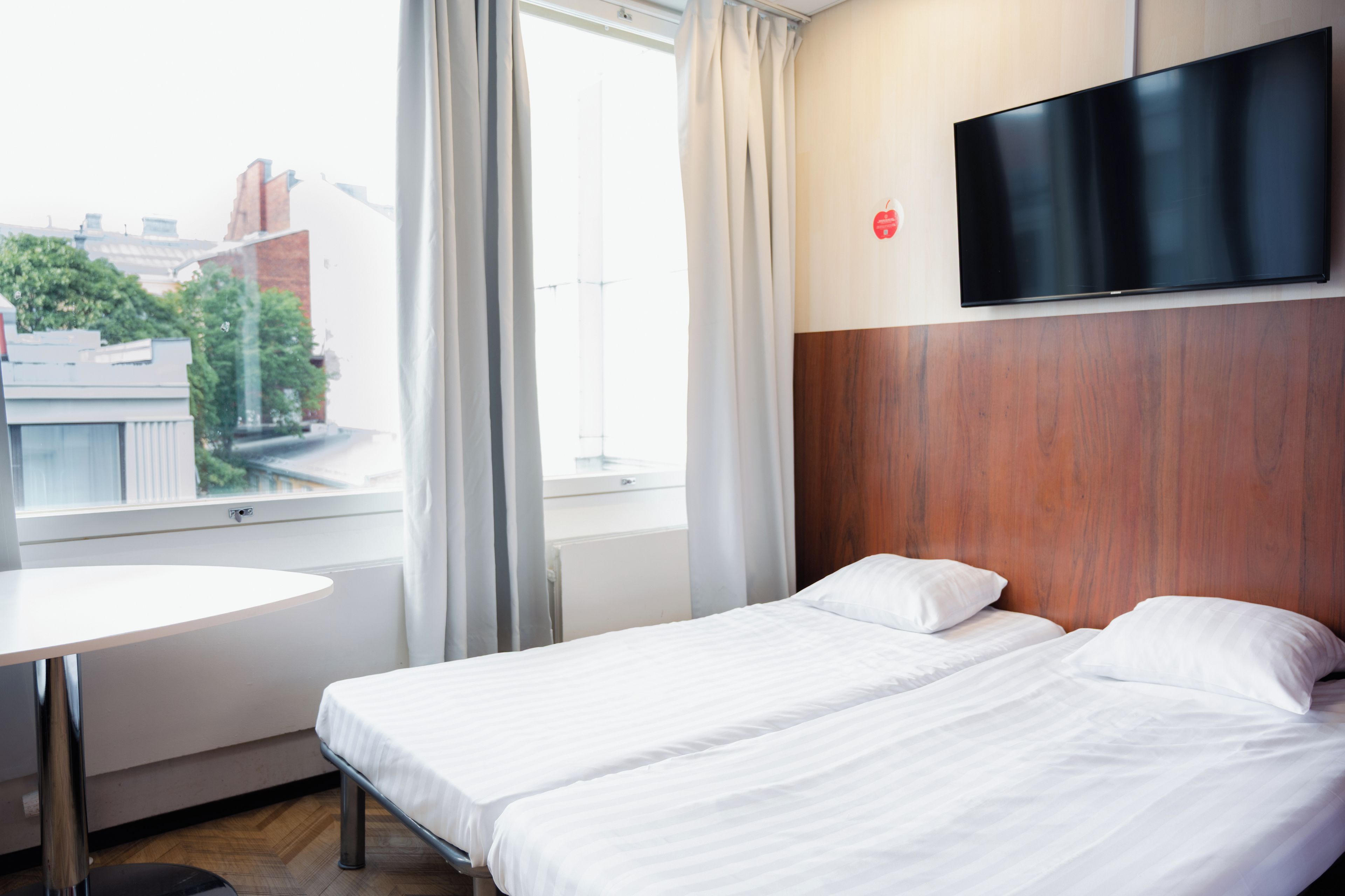 A hotel room with a bed, a TV mounted on the wall, a small table, and a window with curtains.