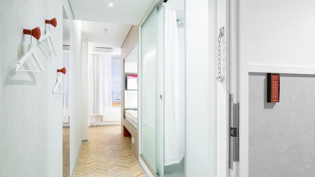 Pasillo de hotel con suelo de madera tipo espiga, cabina de ducha de cristal y cama con ropa blanca al fondo.