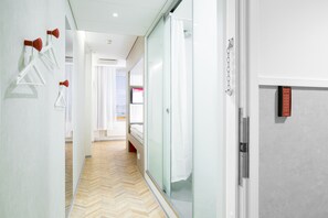 Pasillo de hotel con suelo de madera tipo espiga, cabina de ducha de cristal y cama con ropa blanca al fondo.