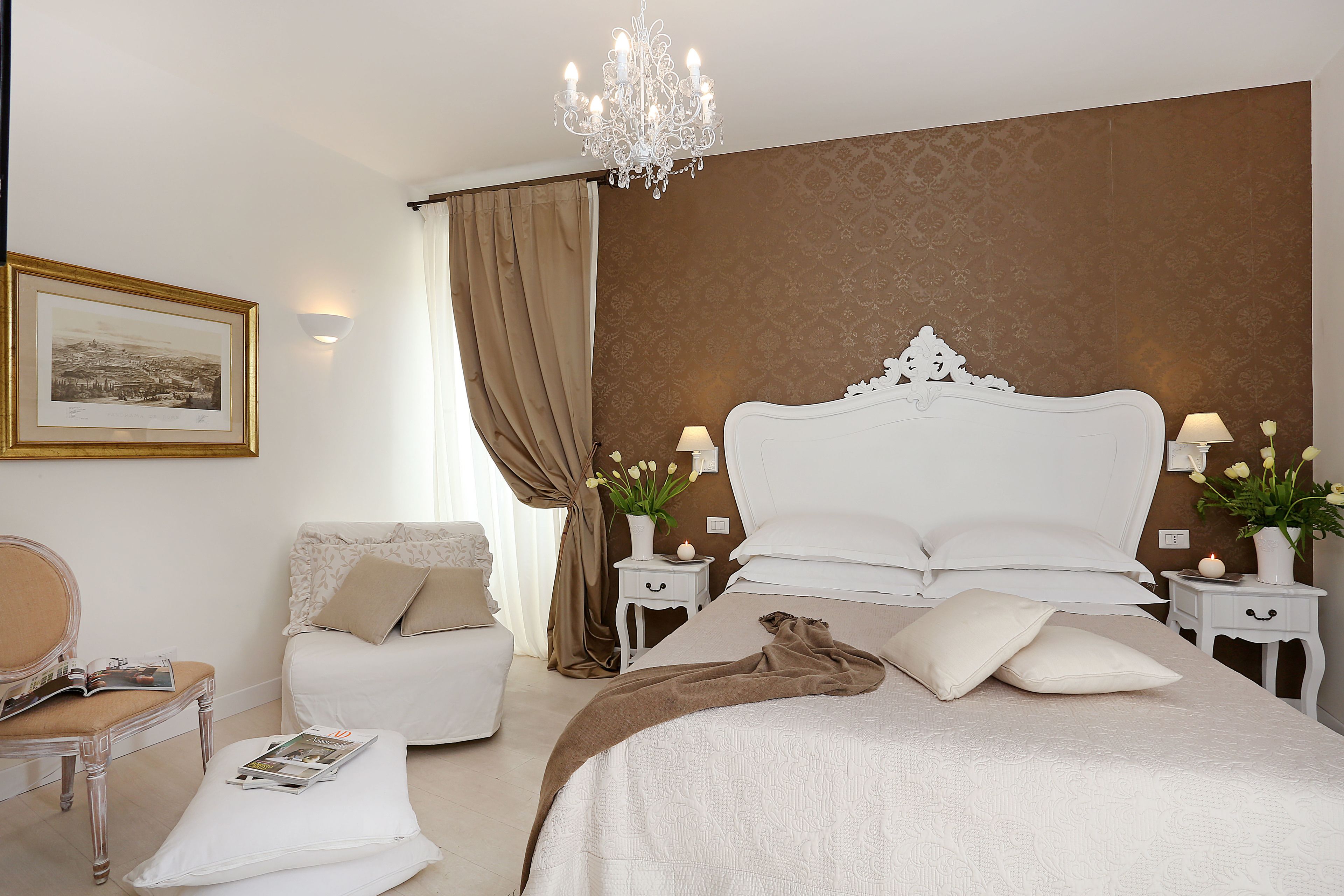 A bedroom with a large bed, two bedside tables with lamps, a chair, a small table with a magazine, and a chandelier.