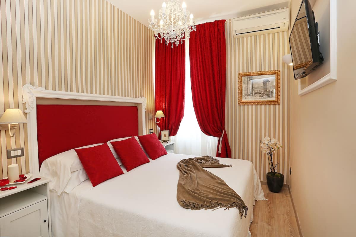 Hotellihuone, jossa on suuri sänky, punaiset verhot, kattokruunu, seinälle asennettu taulutelevisio ja seinällä oleva kehystetty taulu.