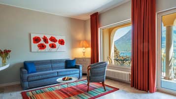 A living room with a sofa, chair, and a table with a plate of fruit. There is a window with red curtains and a view of a mountain.