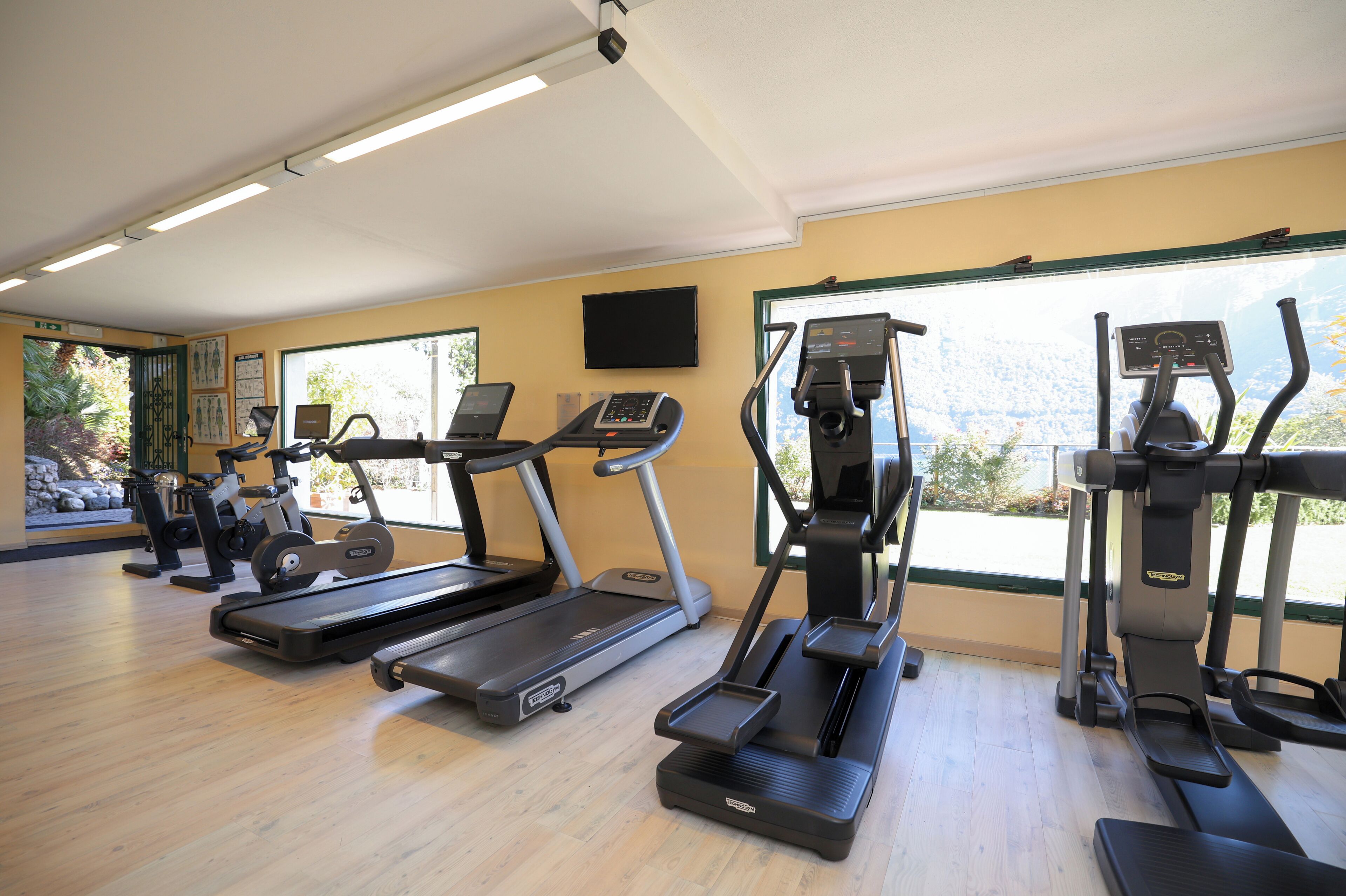 Ein Fitnessstudio mit Laufbändern, Crosstrainern und Ergometer, großen Fenstern mit Aussicht und einem Flachbildfernseher an der Wand.