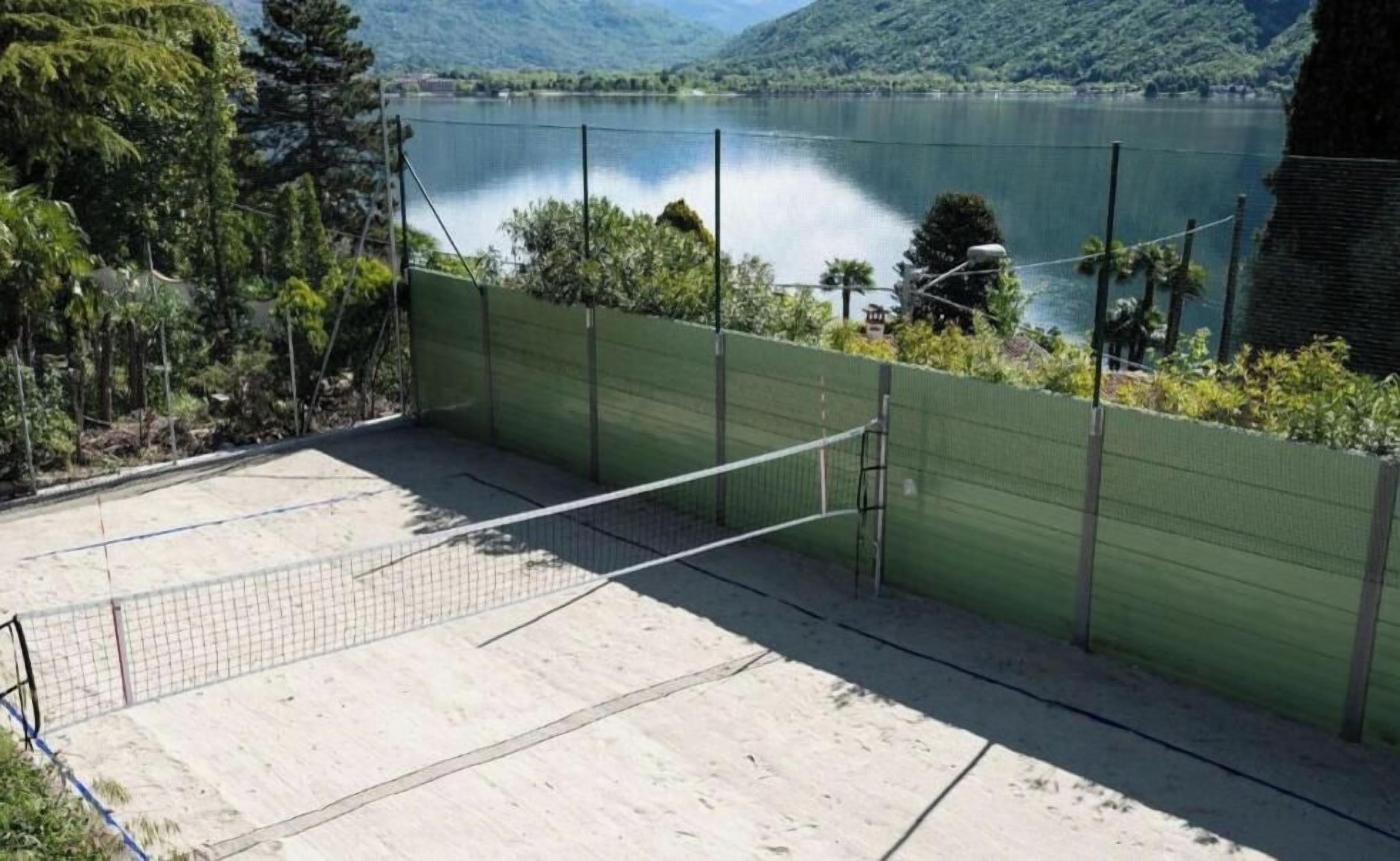 An outdoor tennis court with a net, surrounded by a green fence, and a lake in the background.