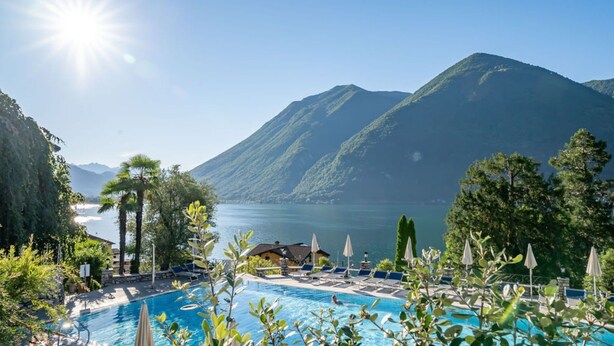 A pool area with lounge chairs and umbrellas, surrounded by mountains and a lake. - Dependence del Parco (Porlezza)