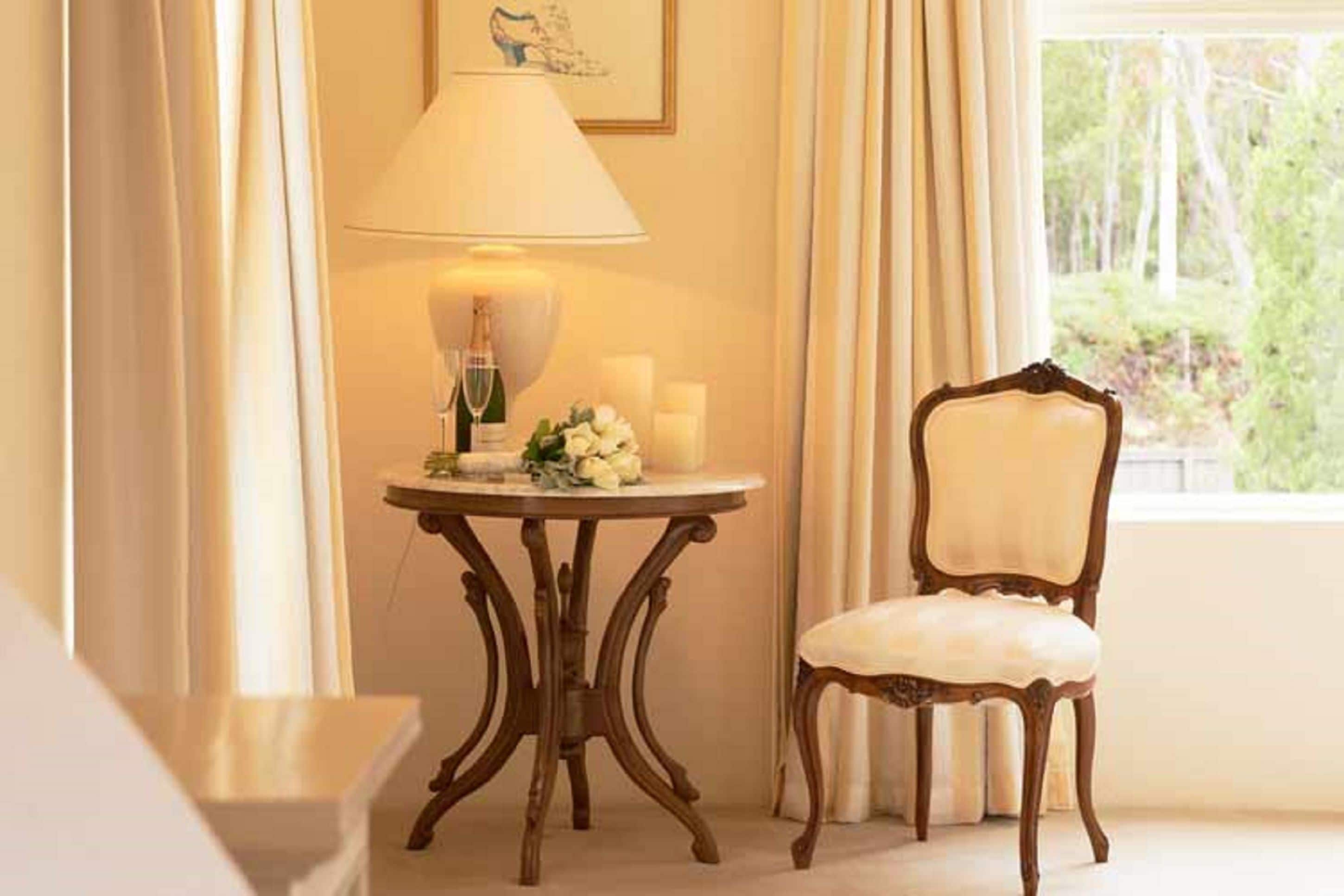 Una habitación con una silla de madera, una mesa con una lámpara, velas y una ventana con cortinas.