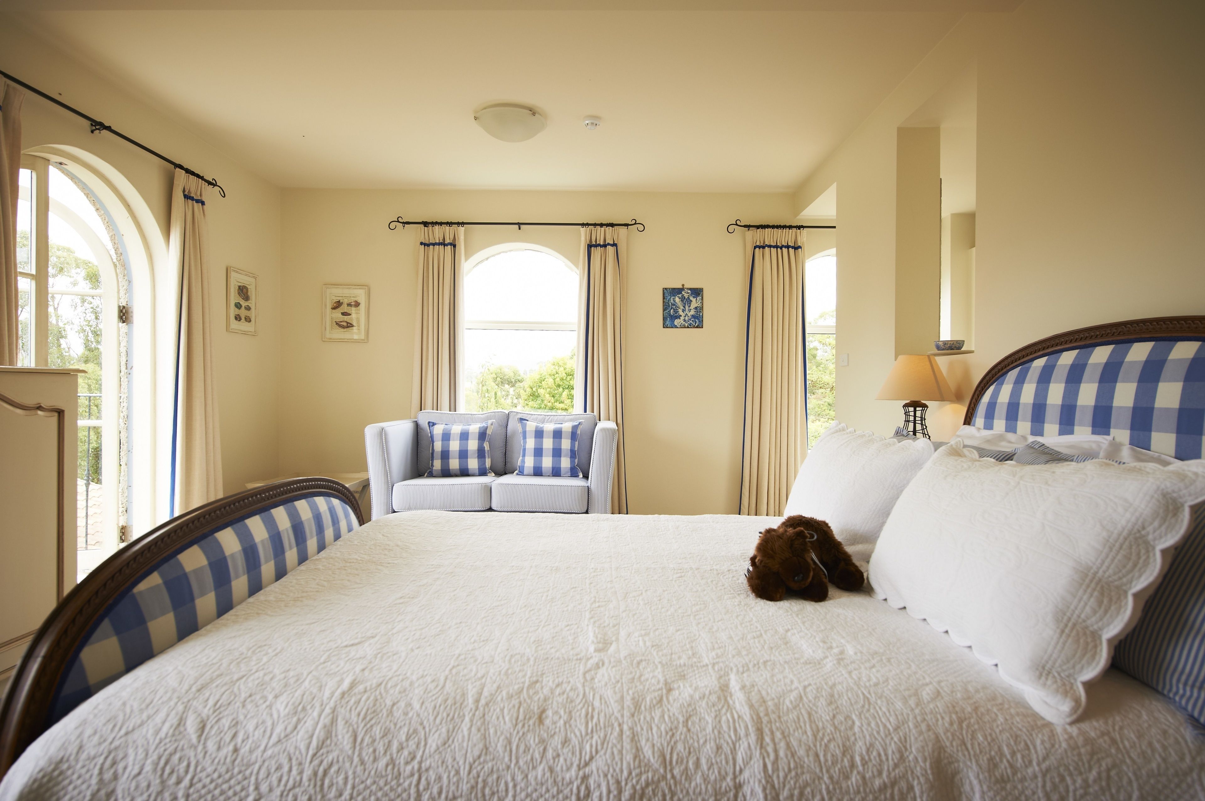 A bedroom with a large bed, two blue and white checkered armchairs, a small table with a lamp, and a window with curtains.