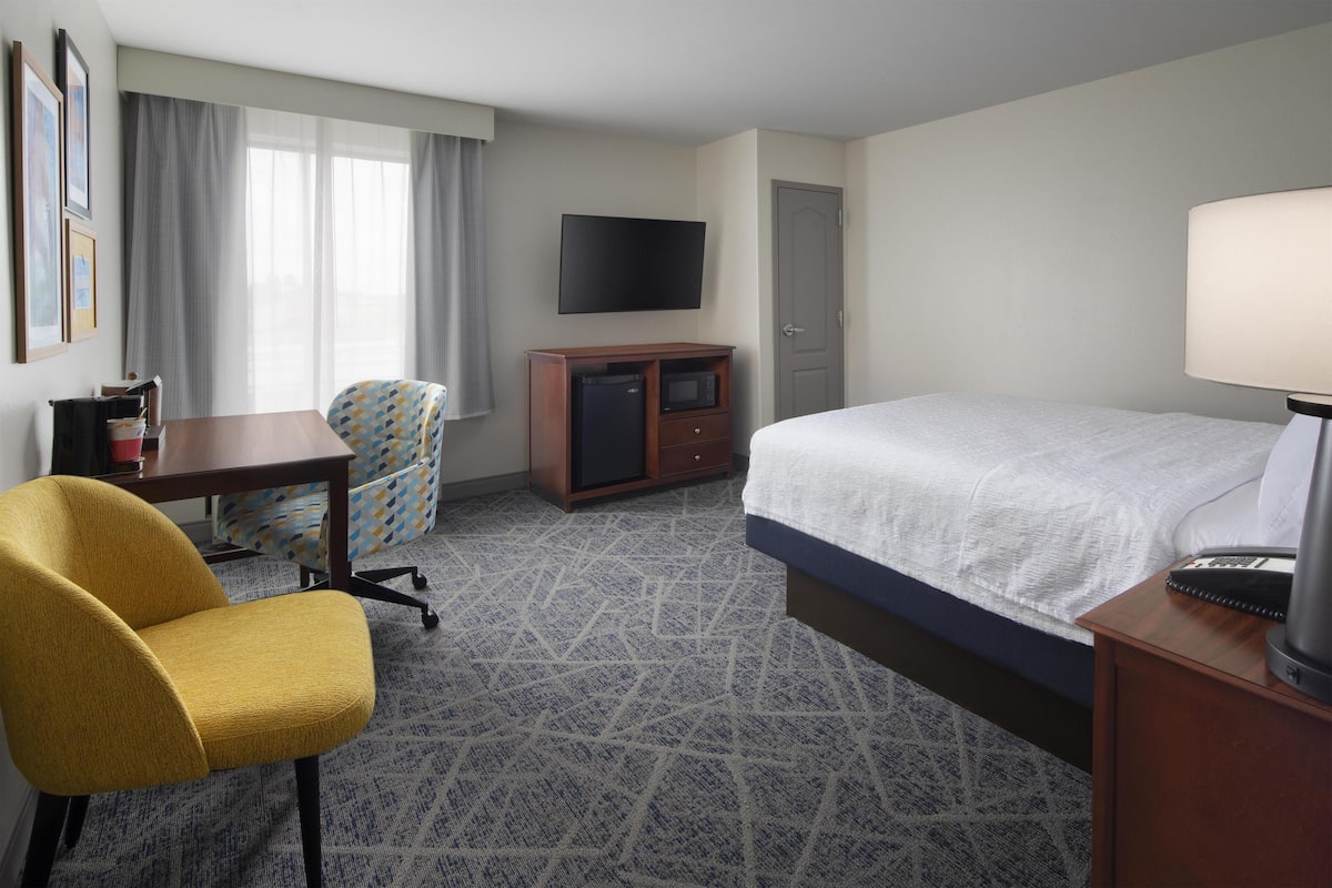 A hotel room with a bed, a desk, a chair, a TV, and a minibar.