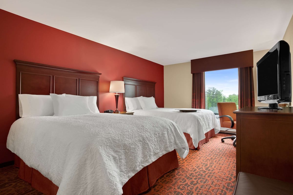 A hotel room with two beds, a desk, a TV, and a window with a view of trees.