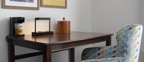 A room with a wooden table, a patterned chair, a coffee machine, and framed pictures on the wall.