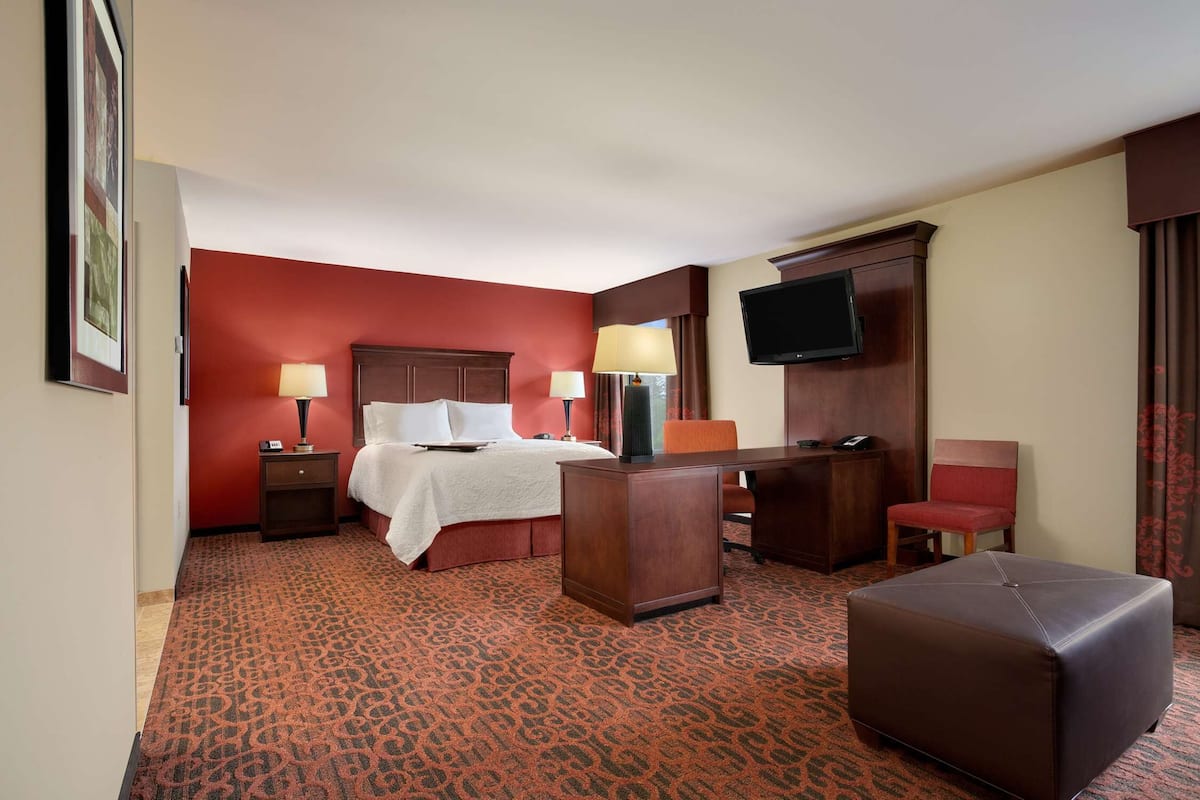 A hotel room with a bed, two bedside tables with lamps, a desk with a chair, a television, and a large ottoman.