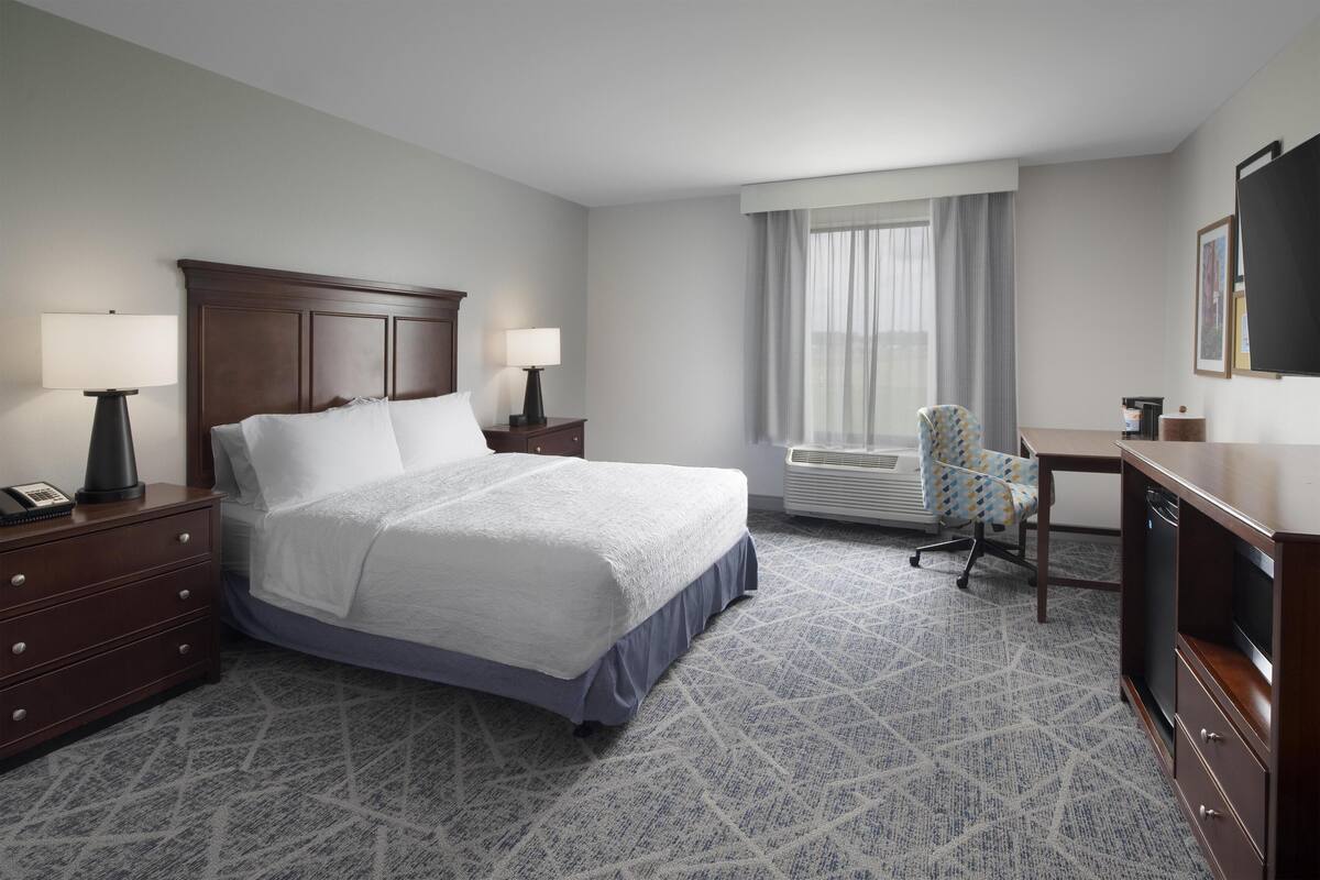 A hotel room with a large bed, two bedside tables with lamps, a desk with a chair, and a large window with curtains.