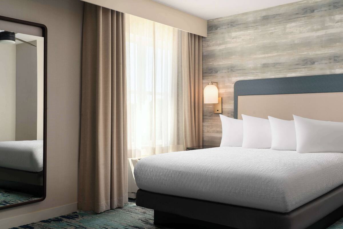 A hotel room with a large bed, a mirror, a wall-mounted light, and a window with curtains.