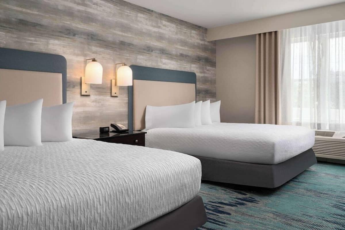 A hotel room with two beds, a wooden textured wall, two wall-mounted lights, a bedside table with a phone, and a large window with curtains.