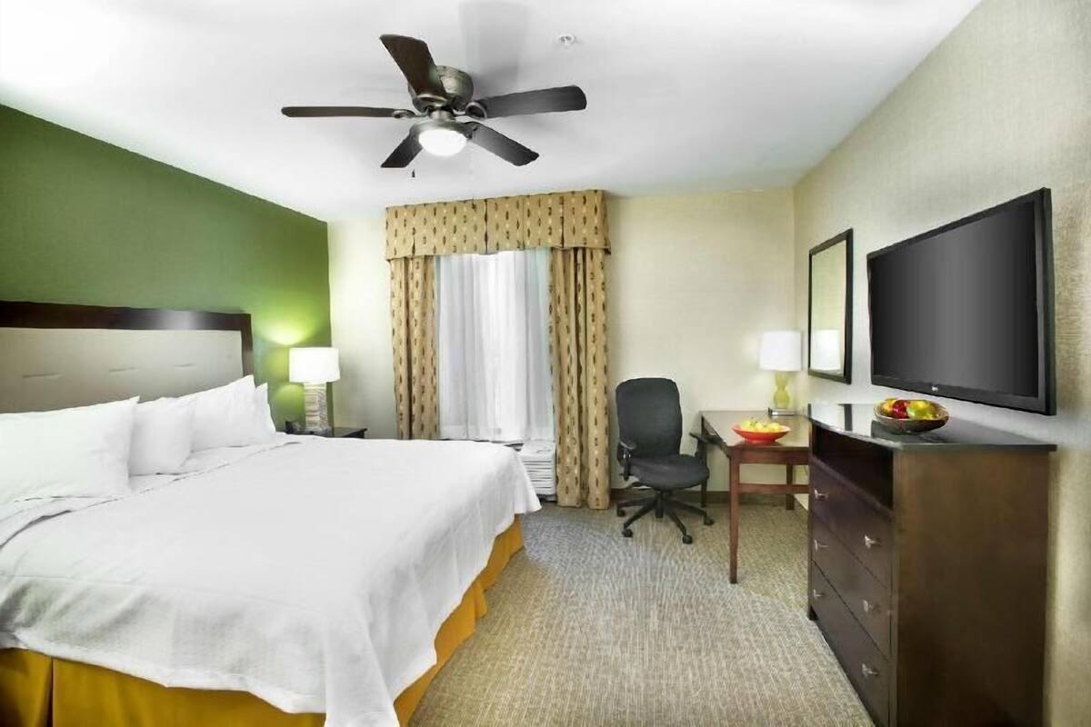 A hotel room with a bed, a desk with a chair, a television, a ceiling fan, and a dresser.