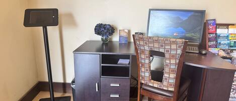 A desk with a computer monitor, a keyboard, and a mouse. There is a chair with a patterned backrest. A small vase with blue flowers is on the desk.