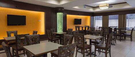 A hotel dining area with multiple tables and chairs, a flat-screen TV mounted on the wall, and a large window with blinds.