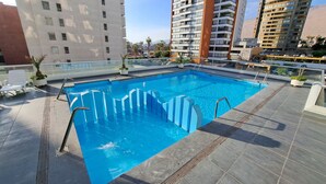 Una piscina en la azotea con agua azul cristalina, rodeada de edificios modernos y palmeras.