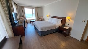 Habitación de hotel con una cama grande, dos lámparas de noche, un escritorio con una silla, una mesita y vistas al océano a través de la ventana.