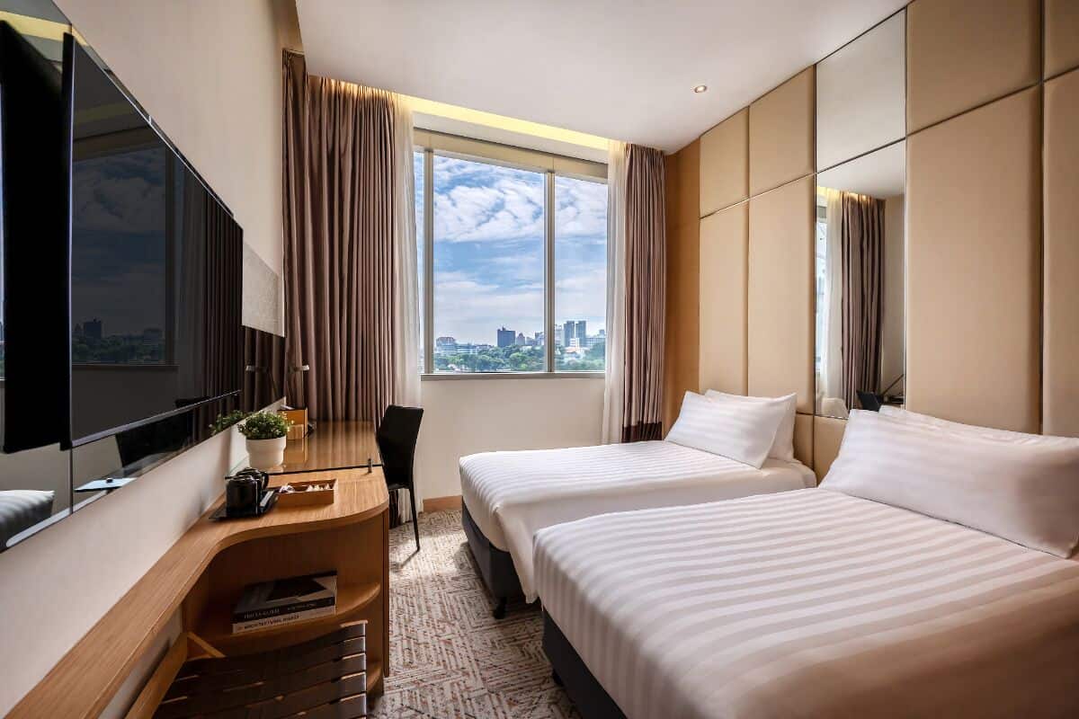 Ein Hotelzimmer mit zwei Betten, einem Schreibtisch, einem Stuhl, einem Fernseher und einem großen Fenster mit Blick auf die Stadt.