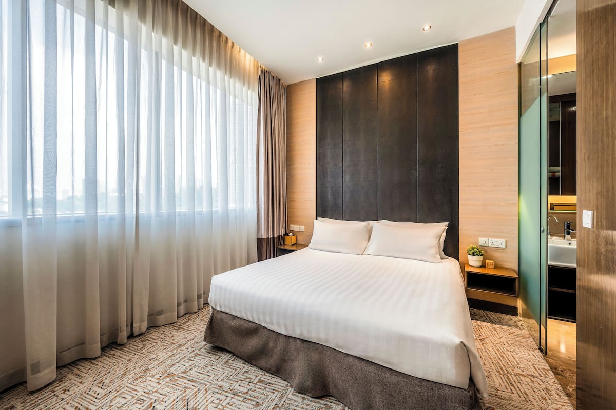 Ein modernes Hotelzimmer mit einem großen Bett, einem Nachttisch, einem Spiegel und Blick auf die Stadt durch große Fenster.