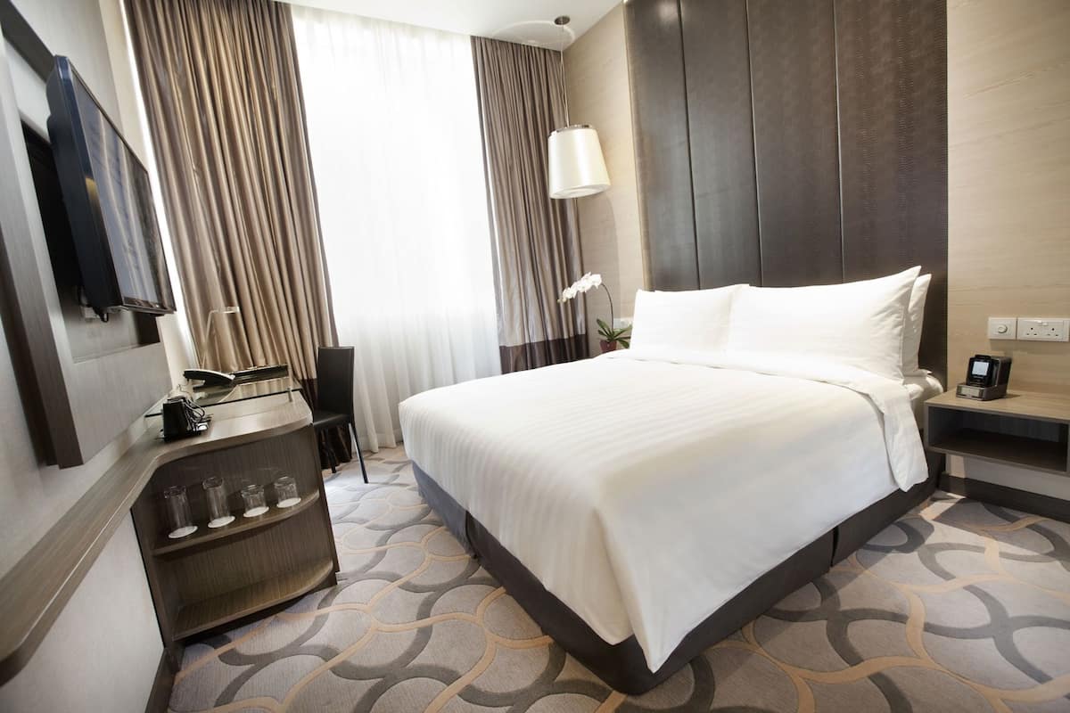 Ein Hotelzimmer mit einem großen Bett, einem Schreibtisch mit Telefon und Lampe, einem an der Wand befestigten Fernseher und einem gemusterten Teppich.