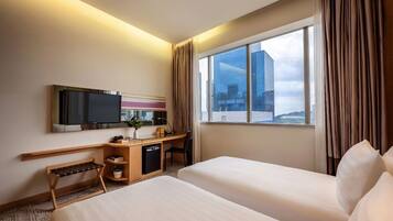 A hotel room with two beds, a desk with a TV, a small seating area, and a window with a city view.