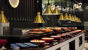Een buffetopstelling met diverse gerechten en kommen, een ventilatie-unit en hangende goudkleurige pendelverlichting.