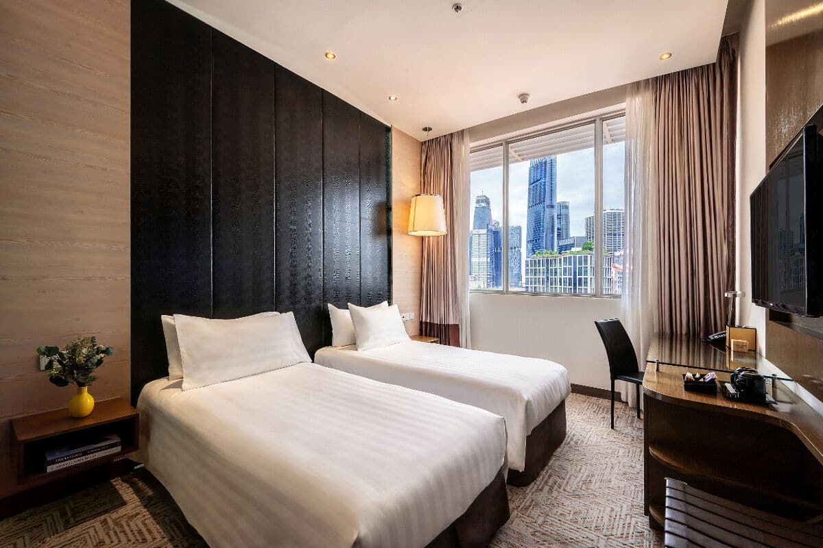 Ein Hotelzimmer mit zwei Betten, einem Schreibtisch, einem Sessel, einem Fernseher und einem großen Fenster mit Blick auf die Stadt.