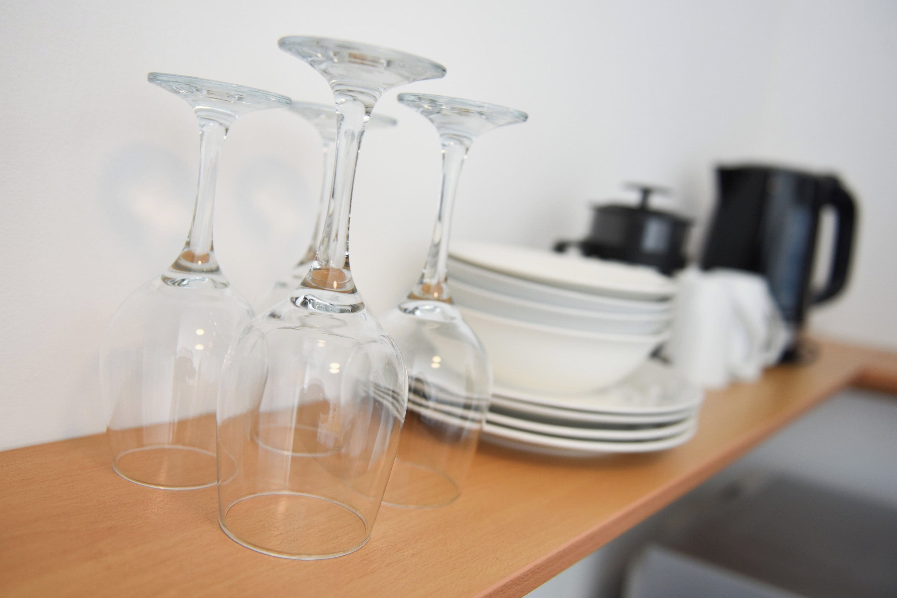 A wooden shelf with three clear wine glasses and a stack of white plates.