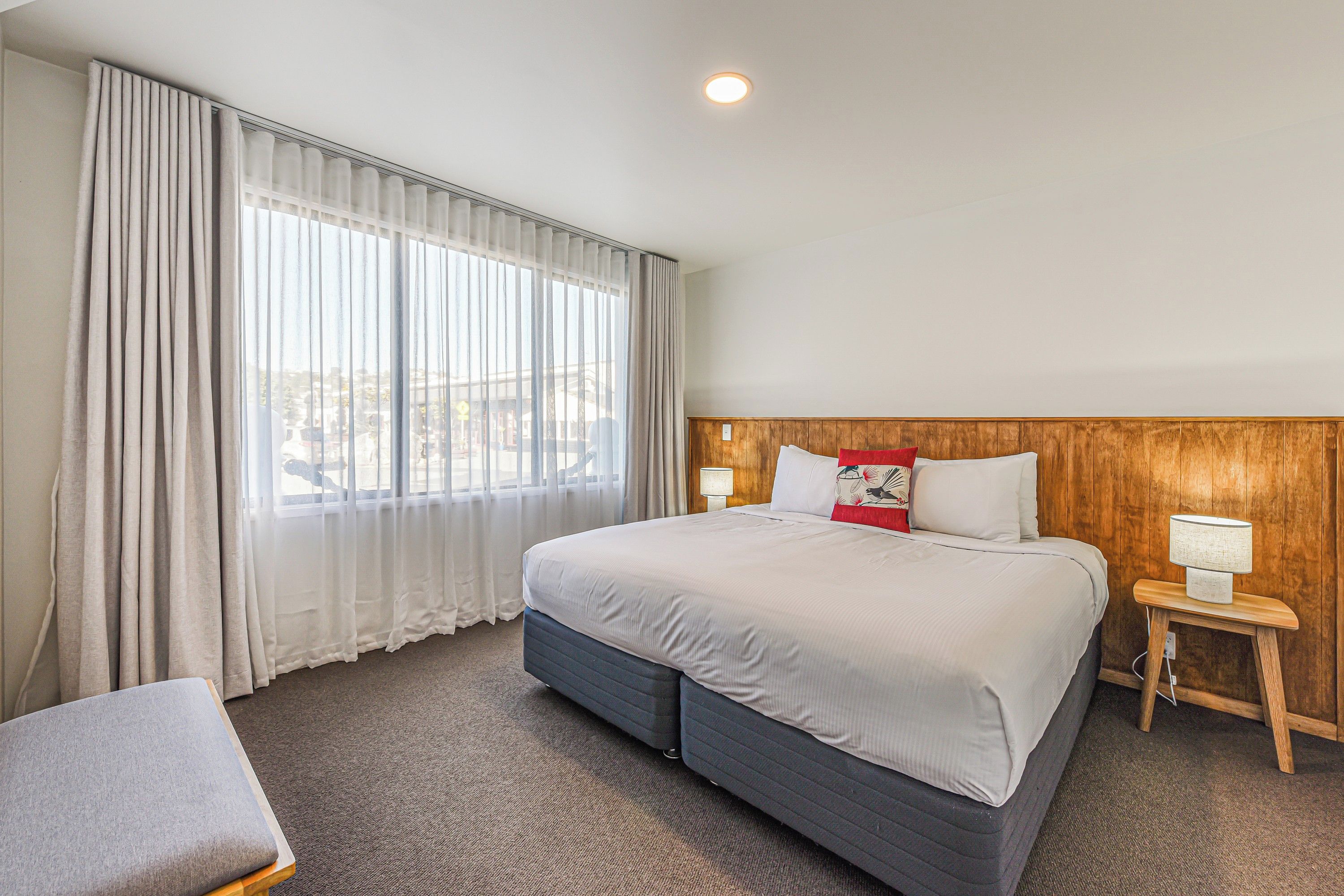 A hotel room with a large bed, a wooden headboard, a bedside table with a lamp, and a view of the outdoors through the window.