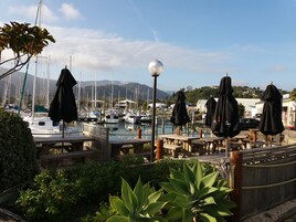 Un puerto deportivo con barcos amarrados, mesas y bancos de madera, y sombrillas.