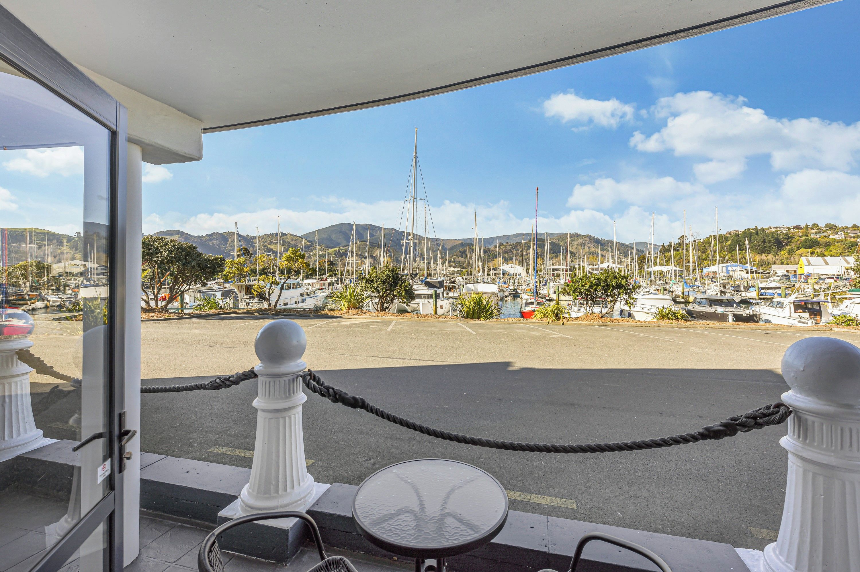 A view from a balcony with a rope barrier, a table, and chairs, overlooking a marina with boats and hills in the background.
