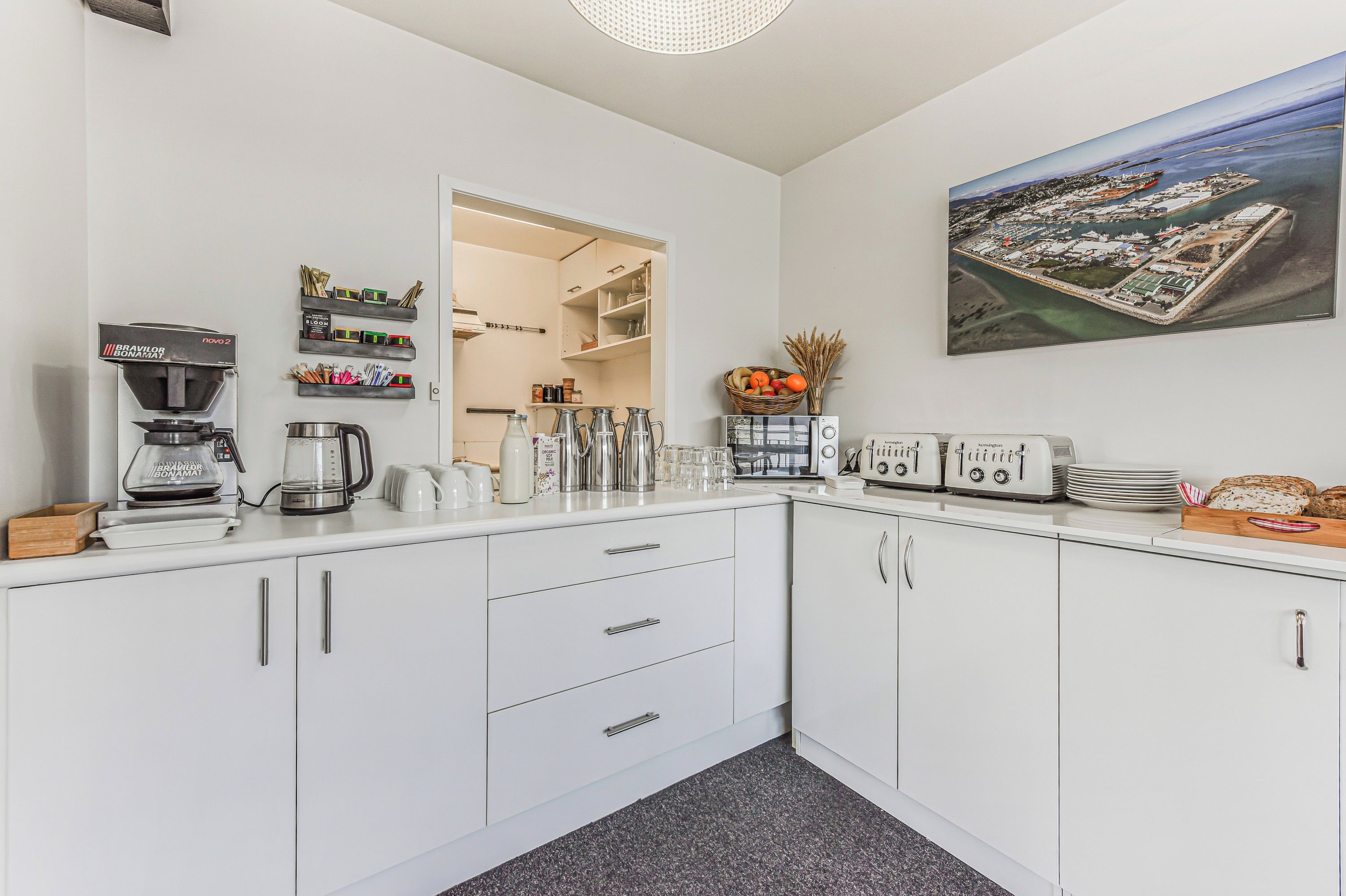 Een moderne keuken met witte kastjes, een koffiezetapparaat, een waterkoker en een broodrooster. Op de muur hangt een lijst met een stadsgezicht.