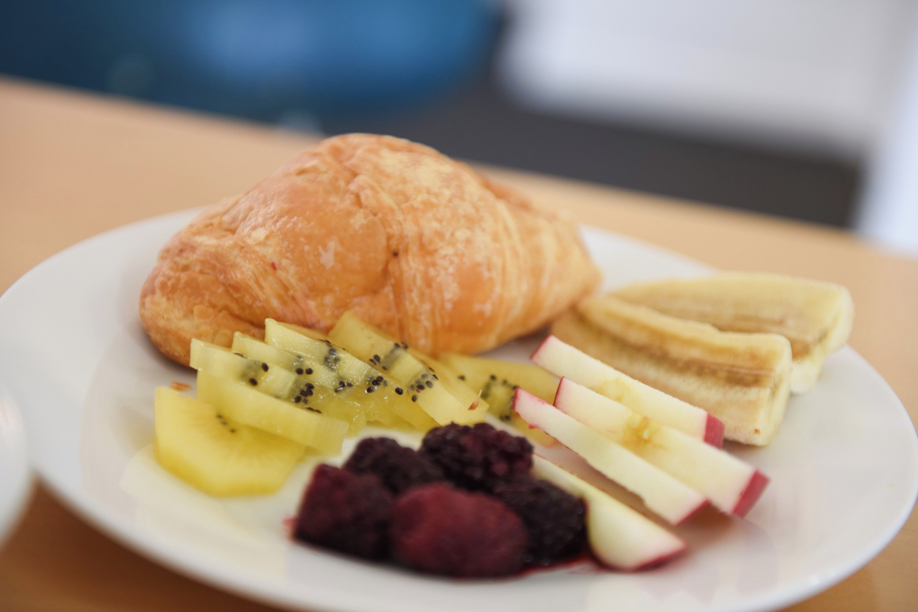 A plate with a croissant, sliced bananas, kiwi, apple, and berries.