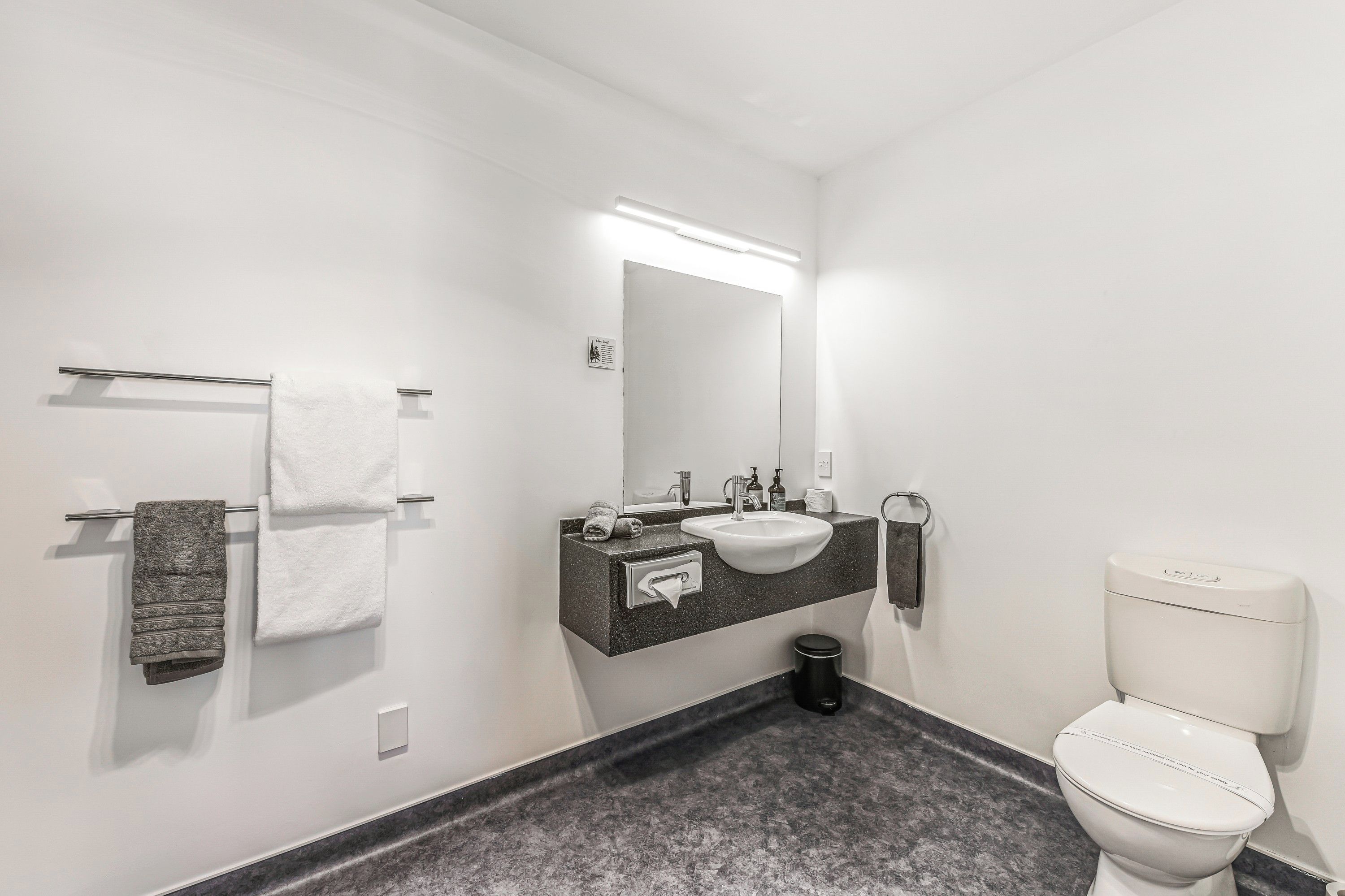 Et moderne badeværelse med et hvidt toilet, en vask med et spejl over og en håndklæderest med to håndklæder.