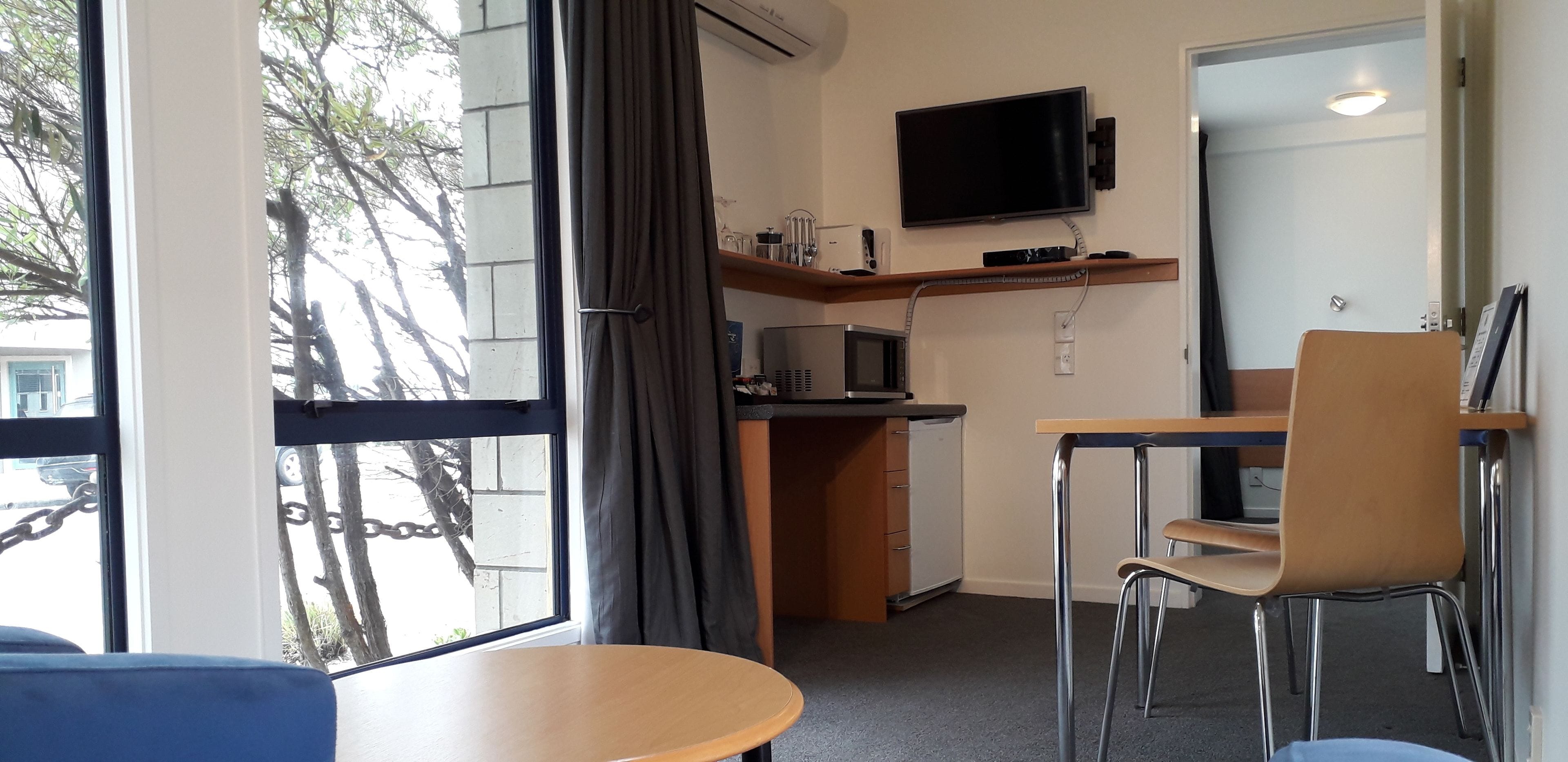A compact hotel room with a kitchenette, a flat-screen TV mounted above the counter, a dining area with a table and chairs, and a large window offering a view of the outside.
