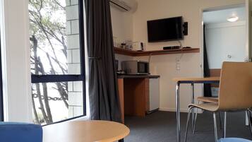 A compact hotel room with a kitchenette, a flat-screen TV mounted above the counter, a dining area with a table and chairs, and a large window offering a view of the outside.