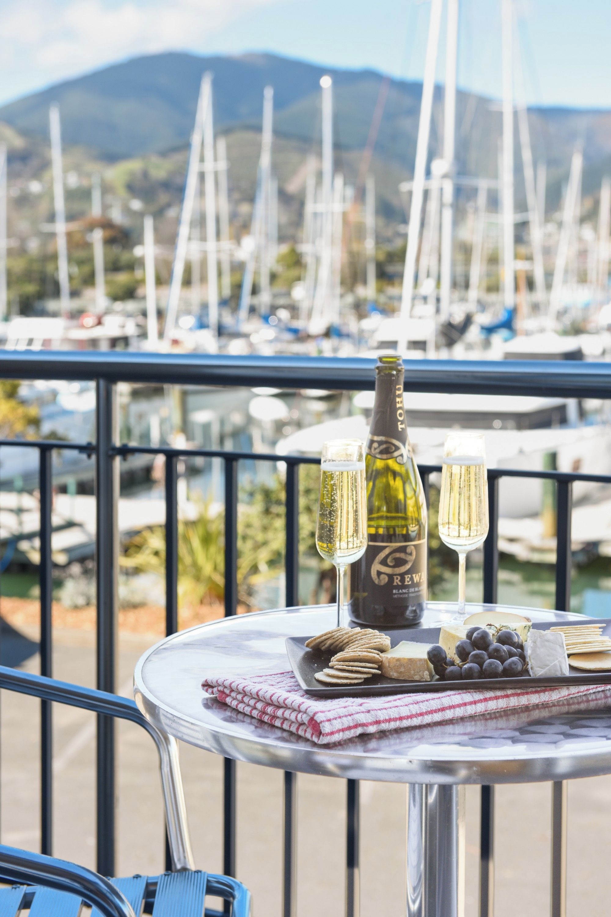 Una mesa con una botella de champán, copas, galletas, queso y uvas en un balcón con vistas a un puerto deportivo lleno de barcos.