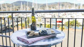 A table with a bottle of wine, glasses, crackers, cheese, and grapes on a balcony overlooking a marina with boats.