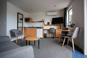 Una sala de estar moderna con un sofá, una mesita redonda y un escritorio con dos sillas. Hay una zona de cocina con microondas, fregadero y refrigerador. Un televisor de pantalla plana está montado en la pared, y hay una planta en maceta sobre el escritorio.